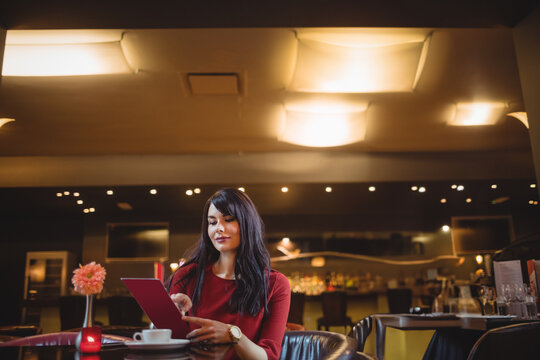 Woman wearing red top sitting at table in restaurant, using tablet near coffee cup, lit candle