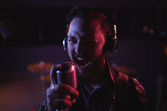 Male singer singing into vintage mic wearing leather jacket and headphones in studio with drums