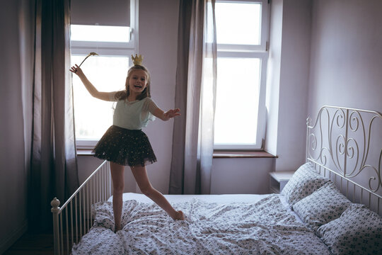 Child girl standing on bed in bedroom wearing sparkly skirt, crown headband and holding toy wand