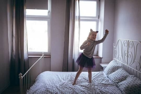 Girl standing on white iron bed in softly lit bedroom with windows and sheer curtains