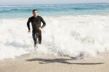 Male athlete running through foamy ocean waves on sandy beach wearing black wetsuit under sunlight
