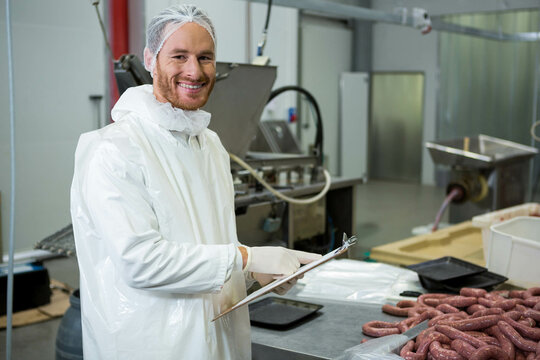 Man inspecting sausage links on steel table at plant in protective coat with clipboard, copy space