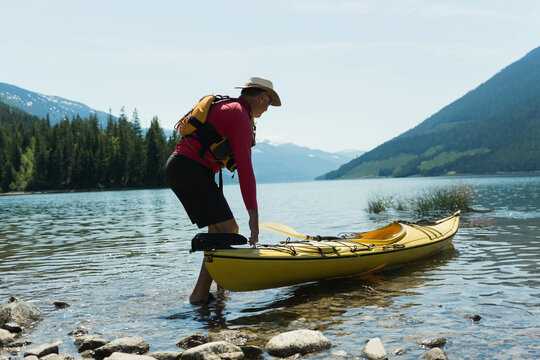 Fototapeta Kayaker placing yellow kayak into clear water on rocky lakeshore with forested hills, snowy peaks