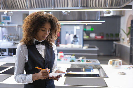 African American server writing on notepad with pencil by induction cooktops in kitchen, copy space