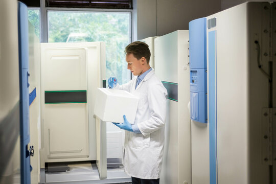 Male lab technician with lab coat and gloves placing samples into cooler box between freezers