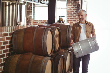 Man holding stainless steel keg inside brewery storage room with oak barrels and tanks, copy space