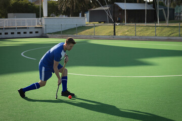 Male field hockey player wearing blue kit gripping stick guiding ball on lined turf, copy space