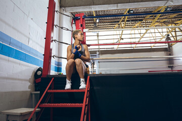 African American female boxer sitting in ring at gym wrapping hand wraps near corner, copy space