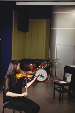Musician holding violin and pressing bow on strings in rehearsal studio near drum kit and keyboard