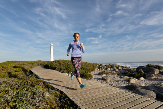 Female runner in sportswear jogging on boardwalk past lighthouse beside rocks and ocean waves
