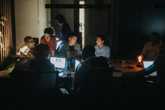 A group of business coworkers engage in a collaborative meeting around a table in a low-light office setting. Laptops and notes are present, emphasizing teamwork and idea sharing.