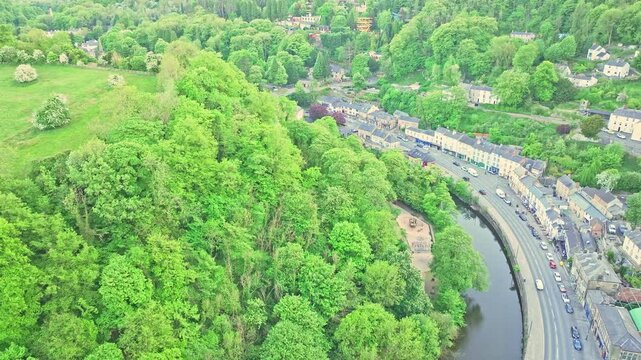 Aerial view captures the thick woodland above the River Derwent&rsquo;s bend, with the Matlock Bath road and historic riverside buildings tracing the curve of the valley in springtime Derbyshire.