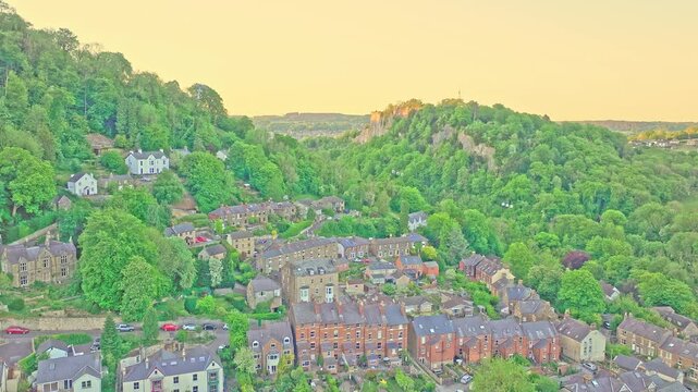 Wide aerial view captures Matlock Bath&rsquo;s rooftops nestled in a green valley beneath High Tor, glowing in golden hour light with dramatic limestone cliff and wooded hills define the Derbyshire scene.