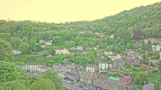 Drone slowly ascends over Matlock Bath, revealing rows of houses climbing the hillside, set within the wooded Derwent Valley. with early spring light highlights the blend of nature and historic charm.
