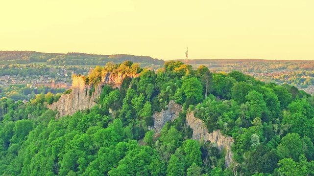 Drone captures High Tor bathed in golden light, a dramatic limestone cliff towering above dense woodland near Matlock Bath in Derbyshire, a landmark popular for climbing, hiking, and scenic views.