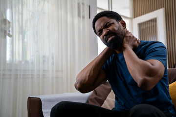 Handsome young African American man sitting on sofa at home living room, holding neck in pain, experiencing neck discomfort during daytime
