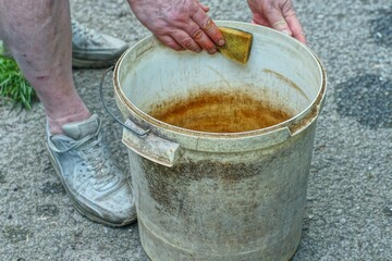 man holding dirty round industrial plastic bucket with iron handle with hands and washing with foam kitchen soft yellow sponge on asphalt in daytime on street