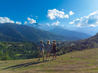 Three tourists are standing on a grassy hilltop, gazing at the stunning panorama of the svaneti mountain range in georgia, under a vibrant blue sky dotted with clouds