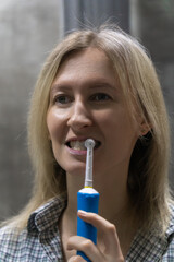 Young woman brushing her teeth with a modern electric toothbrush, maintaining good oral hygiene as part of her daily routine in a domestic bathroom setting. Vertical photo