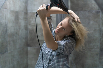 Young blonde woman enjoying her morning routine, drying her hair with a hair dryer in a stylish modern bathroom, surrounded by sleek gray tiles and a sense of comfort