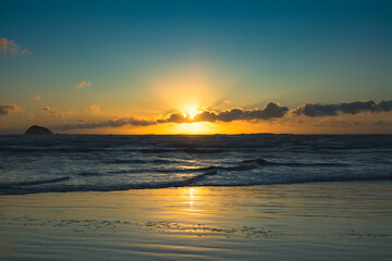 The sun sets behind a veil of clouds over Muriwai Beach, casting golden reflections across the...