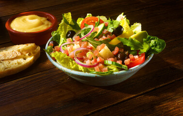 Gemischter Salat auf rustikaler Holzplatte mit Baguette Brotscheibe mit Schinkenwuerfeln und Dip in Terreacotta Schale,