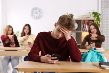 Cyber bullying. Upset teenage boy with smartphone and schoolmates pointing at him in classroom,...