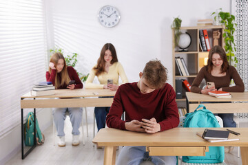 Cyber bullying. Upset teenage boy with smartphone at desk in classroom