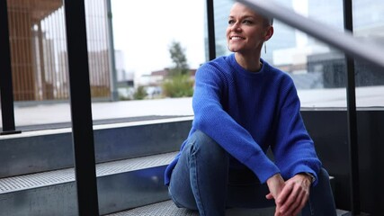 Confident young bald woman in blue sweater and jeans relaxing on modern building stairs, showcasing urban lifestyle with genuine smile and peaceful expression - Powered by Adobe