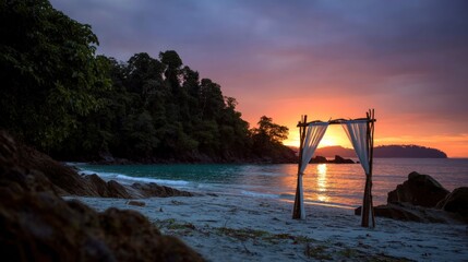 serene beach showcases a decorative arch draped with fabric as the sun sets over calm waters. Lush trees frame the background, creating a peaceful atmosphere in this tropical paradise