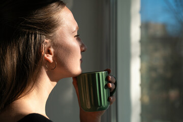 Young woman enjoys a quiet moment, sipping coffee and gazing out the window at the sunny cityscape,...