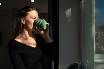 Young woman in a black blouse savoring a tranquil moment while sipping from a green mug by the...