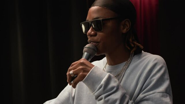 Handheld medium close up shot of confident young Black male rapper in cool shades performing rap into microphone on stage under concert lights