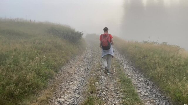 Woman from back with backpack hiking on foggy mountain trail