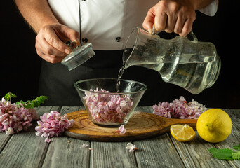 The chef carefully pours water over purple acacia flowers in a glass bowl, surrounded by fresh lemon wedges and herbs, showcasing the calm atmosphere of the kitchen