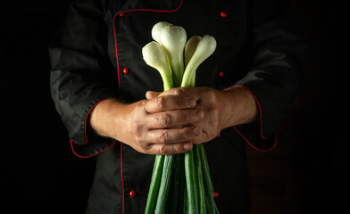 A chef stands in a dimly lit kitchen, holding a bunch of fresh green onions close to their chest,...
