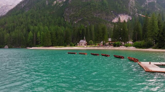 Lago di Braies or Lake Braies crystal blue lake with wooden rowing boats in Italian Dolomites, Lago di Braies chapel and coniferous and broadleaf trees in background, Drone shot