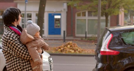 A woman stands on the edge of a street crossing in an urban area while gently holding a baby. In the background, cars are parked neatly and the ground is covered with autumn leaves - Powered by Adobe
