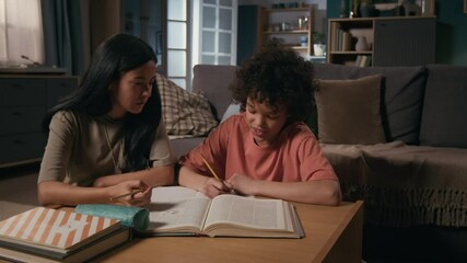 Medium shot of cheerful biracial army soldier mother in military uniform t-shirt sitting with young polyethnic son at home, helping boy with school homework, tenderly stroking hair, patting on head