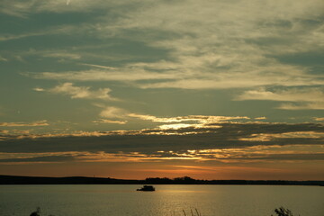 A solitary boat floats on a tranquil lake during a moody sunset. Layers of dark and golden clouds stretch across the sky, casting reflections.