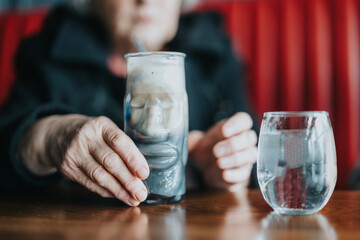 Close-up of a senior womans hands holding a unique glass of iced coffee and water on a wooden surface. Atmosphere of relaxation and reflective moments.