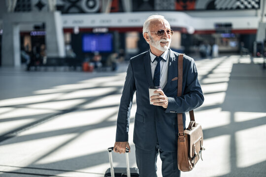 Senior Businessman Walking Through Airport Terminal Holding Coffee and Luggage