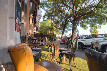 Ornate black metal chairs and round tables on artificial grass, shaded by trees. A tan leather chair is in the foreground, with cars in the background.