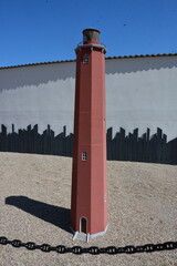 Detailed model of a red brick lighthouse with lantern dome, standing on gravel in bright sunlight....
