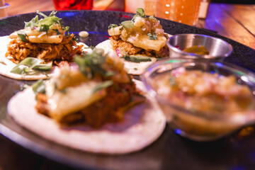 Three gourmet tacos on a black plate with fresh herbs, pineapple, and salsa, accompanied by sauce and salsa bowls on a wooden table.