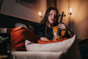 A woman sits on a couch holding a violin in an intimate, cozy ambient setting. The mood conveys a...