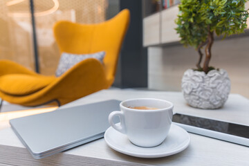 A cozy interior with a coffee cup, laptop, and tablet on a wooden table. A mustard yellow armchair, plant, and bookshelves complete the setting.