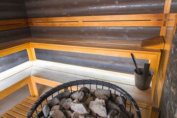 Luxury sauna featuring light wood benches, dark wood paneling, and a central heating element with stones, bucket, and ladle in a sleek design.