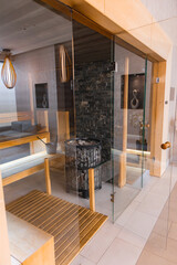 Luxurious spa interior featuring a glass walled sauna, light wood details, stone wall with central heater, tiled floor, and sleek lighting fixtures.