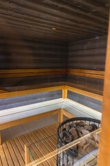 Interior of a modern sauna with tiered wooden seating, polished wood paneling, and a central heating element with sauna stones in a metal frame.
